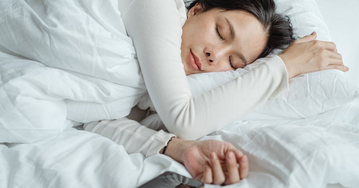 Calm Asian female wearing white pajama sleeping in comfortable bed with white sheets near modern mobile phone in morning